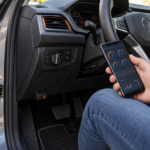 Interfejs OBD2 podłączony pod deską rozdzielczą i smartfon z parametrami diagnostycznymi w dłoni kierowcy zaparkowanego auta.